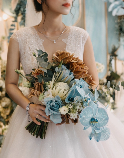 Trong ngày vui, cô dâu diện váy cưới dáng xòe, tay ren và lựa chọn hoa cầm tay gồm có: thảo đường hoàng đế, lan hồ điệp, lá bạc, hoa hồng Ecuador màu nâu cà phê (toffee) theo tông màu tiệc.